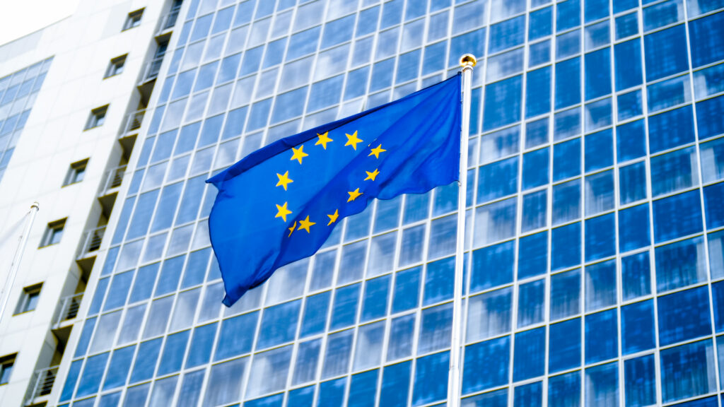 Image of EU flag fluttering on wind against high business office building made of concerete and glass. Concept of ecenomics, development, government and politics